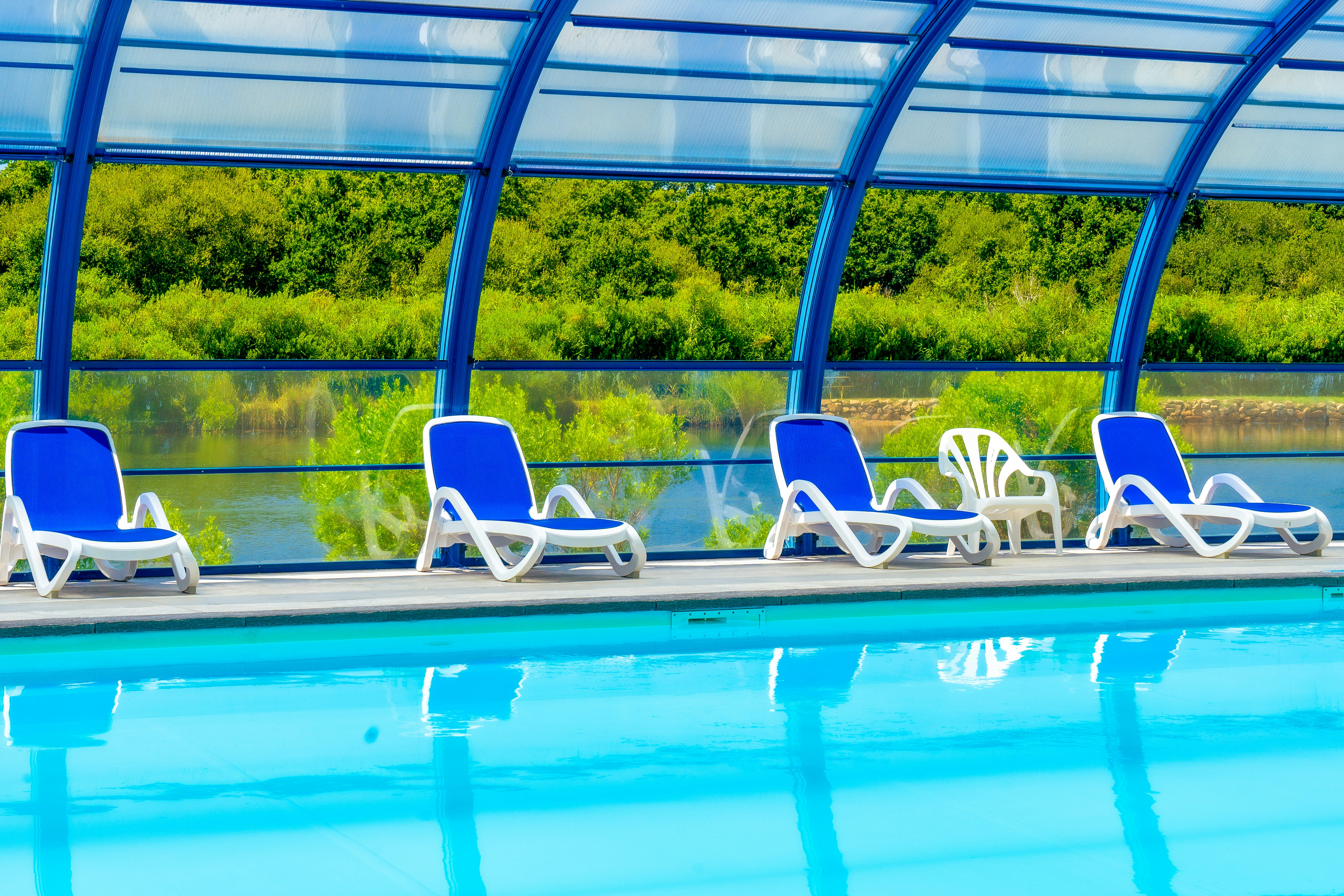 Blaue Pool-Liegen unter einer transparenten Überdachung am Schwimmbecken, im Hintergrund grünes Ufer und Wasser.