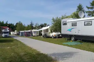 Wohnwagen auf einem Campingplatz entlang eines gepflasterten Wegs, daneben Zelte und weitere Fahrzeuge zwischen Bäumen.