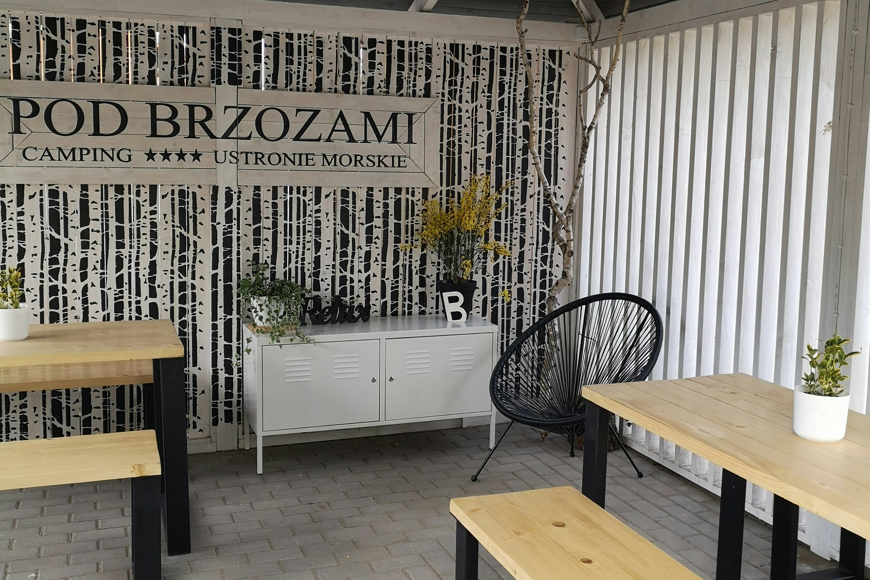 Holz-Tisch und schwarze Gartenstühle auf der Terrasse, daneben ein weißer Schrank und Deko vor dem Camping-Banner „Pod Brzozami“.