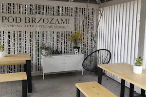 Holz-Tisch und schwarze Gartenstühle auf der Terrasse, daneben ein weißer Schrank und Deko vor dem Camping-Banner „Pod Brzozami“.