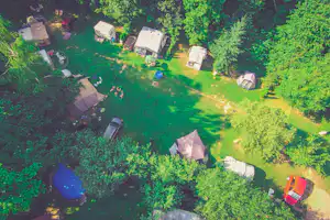 Luftaufnahme eines Campingplatzes mit Zelten, Wohnwagen und mehreren Stellplätzen im Grünen, umgeben von Bäumen und Wegen.