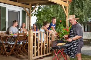 Familie beim Grillen vor einer Holzveranda am Campinghaus, mit Esstisch, Stühlen und grünen Pflanzen im Hintergrund.