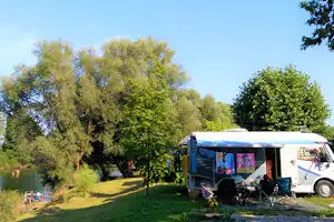 Wohnmobil am Flussufer im Grünen mit Markise, Campingstühlen und Tisch auf dem Stellplatz zwischen Bäumen.