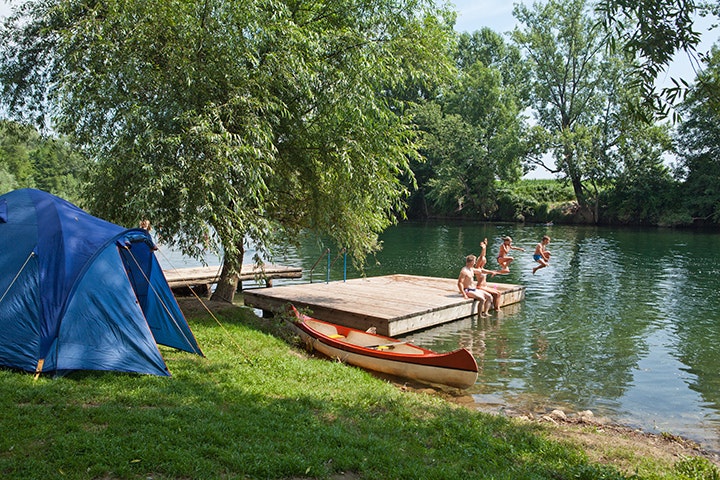Blaues Zelt am Seeufer, Holzsteg mit Ruderbooten und Personen im Wasser, umgeben von Bäumen.