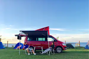 Rotes Wohnmobil mit ausgeklappter Markise, Campingstühle und Zelt am Grasplatz direkt am Meer.