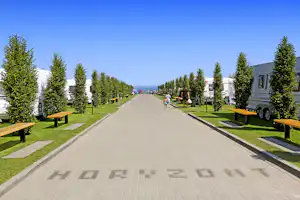 Camping-Allee mit gepflastertem Weg, Sitzbänken und Hecken zwischen Stellplätzen, im Hintergrund Bäume und das Meer.