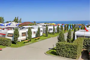 Reihe von Wohnwagen auf einem Campingplatz, gepflegter Rasen mit Hecken, Weg und Meerblick unter klarem Himmel.