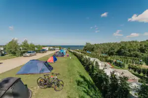 Zelte auf einem Campingplatz am Hang, Fahrräder am Weg, Wohnwagen und Autos in der Nähe, Blick auf das Meer.