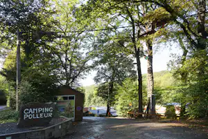 Eingang zum Camping Poleur mit Schild, Schotterweg und Bäumen im Vordergrund, dahinter Wohnwagen und Wiese am Hang.