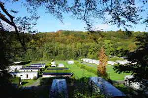 Campinganlage im grünen Tal mit Rasenflächen, Wohnwagen und Zelten, umgeben von Bäumen und Hügeln.