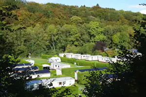 Campingplatz mit Wohnwagen auf grünen Stellplätzen, umgeben von Bäumen und Hügeln im Tal.