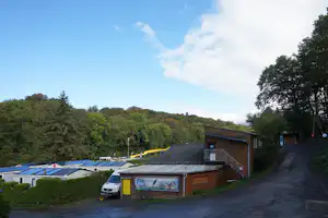 Kleines Haus am Campingplatz mit gelber Tür, davor ein Auto, im Hintergrund Stellplätze und Bäume.
