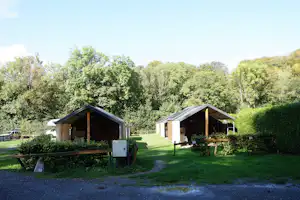 Zwei Camping-Bungalows mit Holzfassaden und kleinen Veranden stehen im Grünen, mit Wegen und Rasenflächen im Hintergrund.