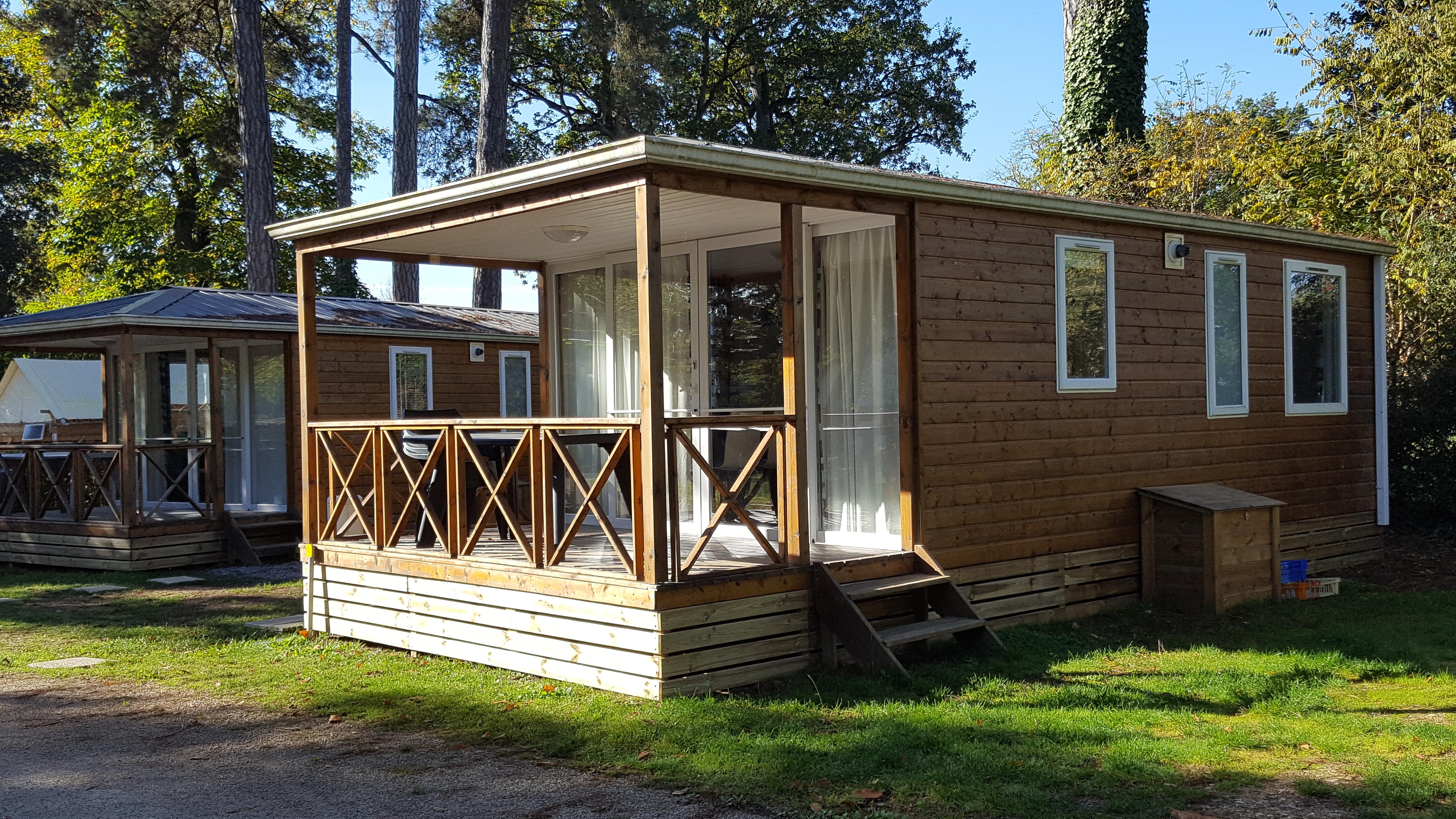 Holzbungalow mit Veranda und großen Fenstern, daneben ein weiterer Bungalow im Grünen zwischen hohen Bäumen.