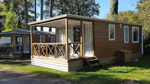 Holz-Bungalow mit Veranda, großen Fenstern und Vorhang, daneben ein weiterer Bungalow im Grünen unter hohen Bäumen.