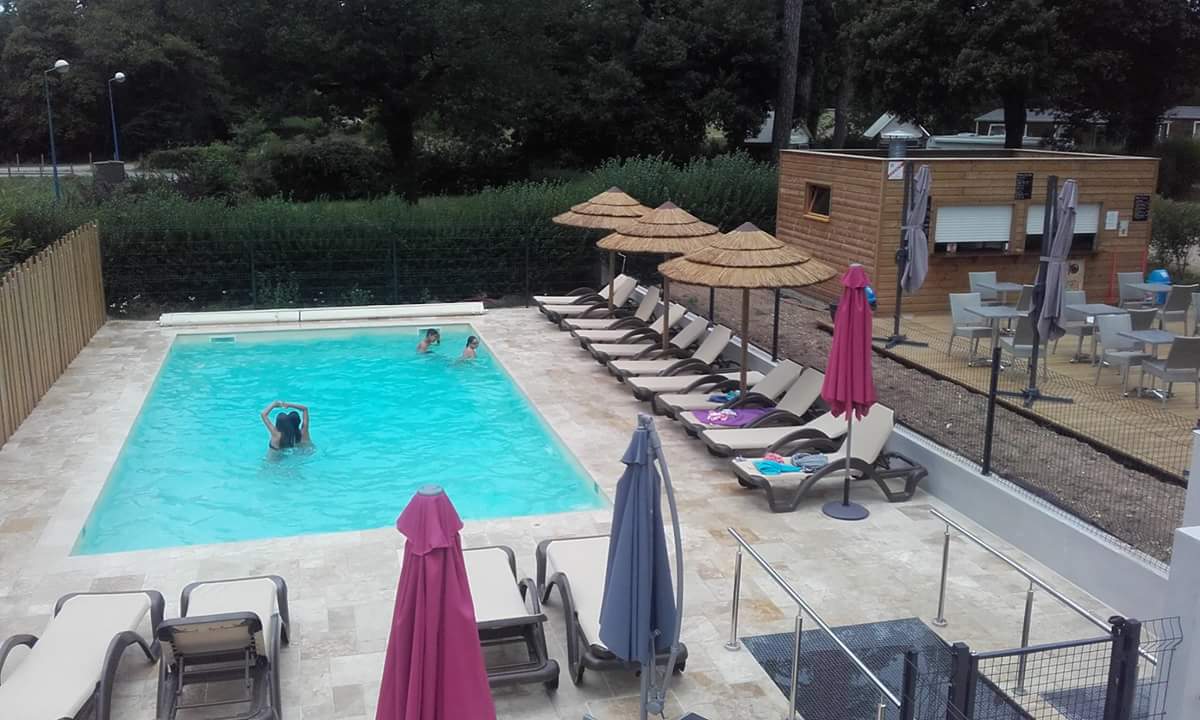 Schwimmbecken mit Wasser, Liegen und Sonnenschirmen am Campingplatz, daneben ein Holzgebäude und ein Zaun mit Stufen.