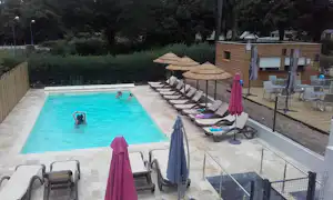 Schwimmbecken mit Wasser, Liegen und Sonnenschirmen am Campingplatz, daneben ein Holzgebäude und ein Zaun mit Stufen.