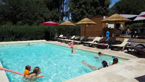 Schwimmbecken mit türkisfarbenem Wasser, Kinder planschen, daneben Liegen und Sonnenschirme vor einem Holzgebäude.