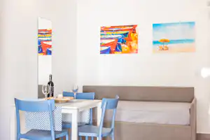 Wohnbereich mit Sofa und Esstisch, blauen Stühlen, Weinflasche und Gläsern sowie Wandbildern an der weißen Wand.
