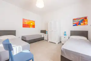 Schlafzimmer mit zwei Betten, weißem Schrank mit Läden, Nachttischen und bunten Wandbildern an der hellen Wand.