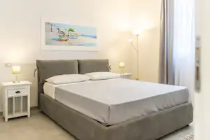 Doppelbett im Schlafzimmer mit weißen Laken, grauem Kopfteil, Nachttischen und Lampen sowie Wandbild und Vorhang am Fenster.