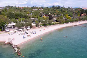 Campingplatz am See mit Sandstrand, Liegen und Blick auf das Ufer sowie Häuser und Bäume am Ort.