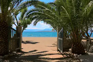 Blick durch ein offenes Tor auf den Sandstrand, Palmen und Meer mit Bergen im Hintergrund.