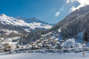 Winterliches Bergdorf mit vielen Häusern, verschneiten Wegen und Skigelände, umgeben von Tannen und Bergen.