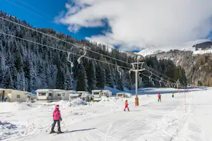 Skigebiet mit Sessellift über verschneiten Pisten, im Vordergrund Campingwagen und Wohnmobile am Rand des Waldes.