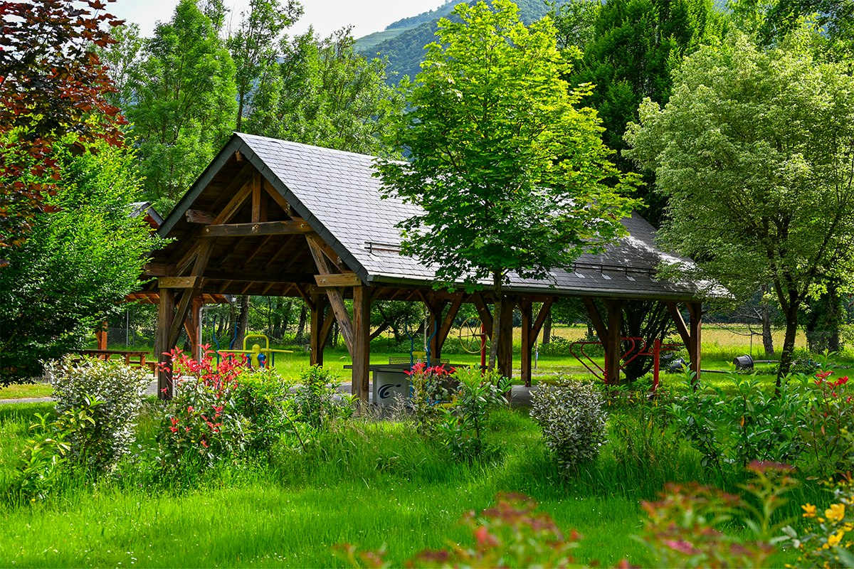Holzüberdachter Pavillon mit Sitzbereich auf einem Campingplatz, umgeben von grünen Bäumen und gepflegten Blumenbeeten.