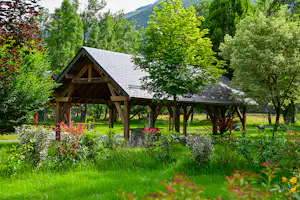 Holzüberdachter Pavillon mit Sitzbereich auf einem Campingplatz, umgeben von grünen Bäumen und gepflegten Blumenbeeten.