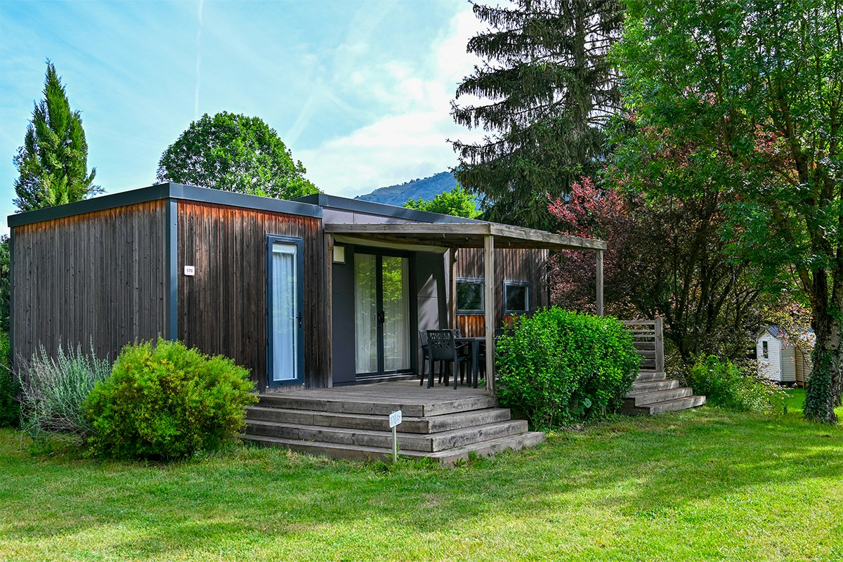 Modernes Campinghaus mit Holzveranda und Stufen, Sitzgruppe auf der Terrasse, umgeben von Büschen und Bäumen im Garten.
