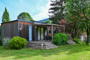 Modernes Campinghaus mit Holzveranda und Stufen, Sitzgruppe auf der Terrasse, umgeben von Büschen und Bäumen im Garten.