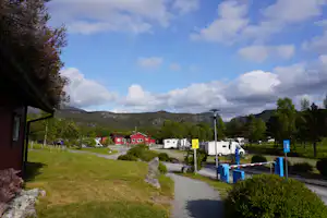 Campingplatz mit Wohnwagen und Stellplätzen, gepflastertem Weg, Bäumen und Wiesen; im Hintergrund rote Häuser und Hügel.