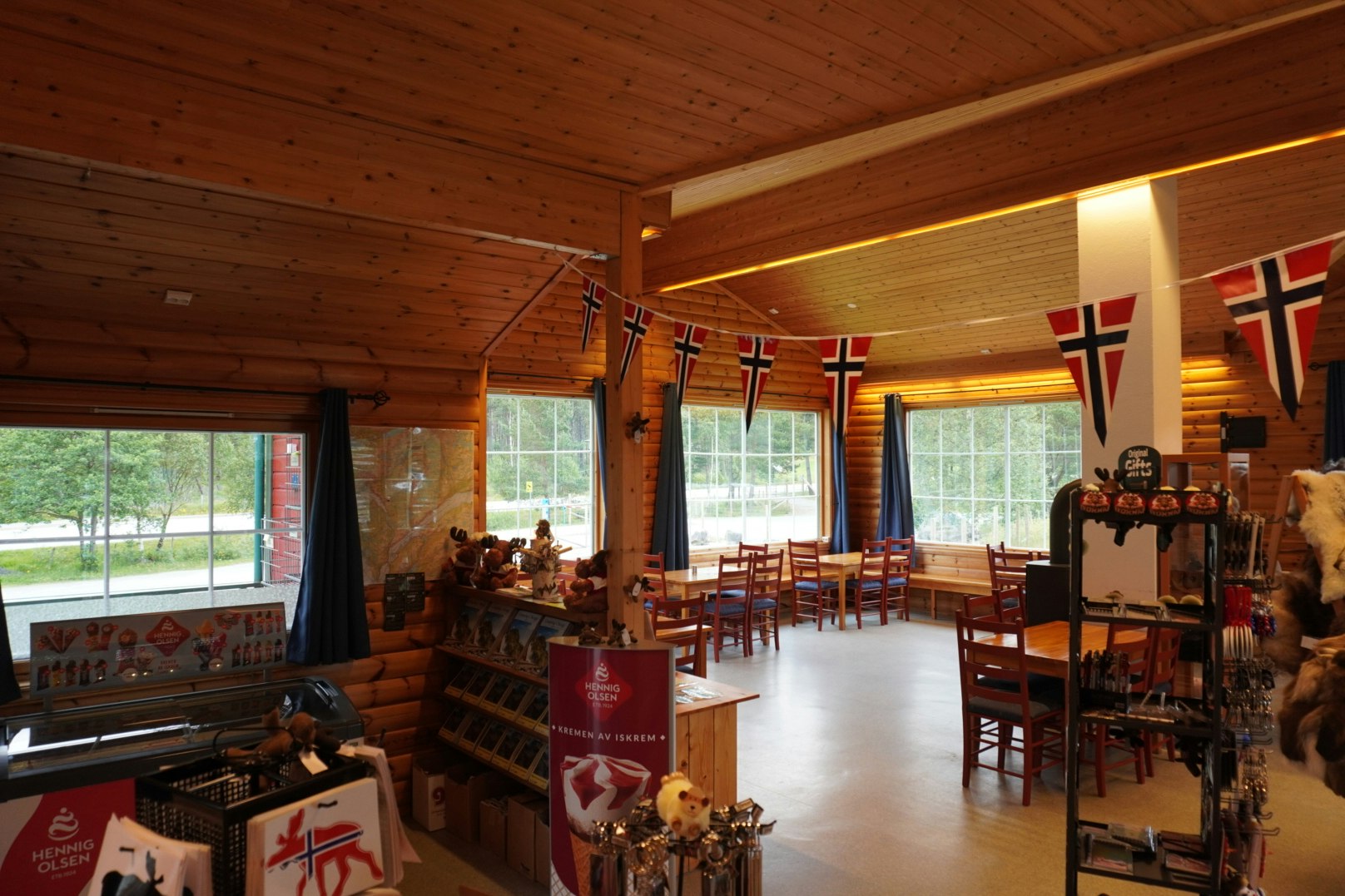 Innenraum einer Blockhütte im Campingbereich mit Holzwänden, Tischen, Stühlen und mehreren Flaggen an der Wand.