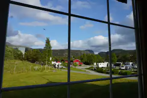 Blick durch ein Fenster auf einen Campingplatz mit Rasen, Wohnwagen, Autos und roten Häusern inmitten von grünen Hügeln.