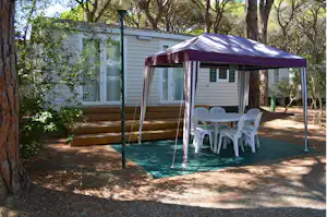 Wohnwagen mit überdachtem Sitzbereich, weißen Kunststoffstühlen und Tisch vor einer Holzterrasse, umgeben von Bäumen und Grün.