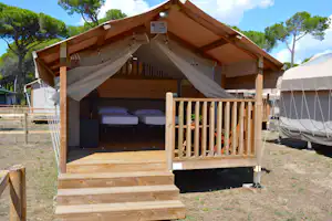 Offenes Campingzelt mit Holzveranda, zwei Betten und Vorhängen, daneben ein weiteres Zelt auf dem Stellplatz.