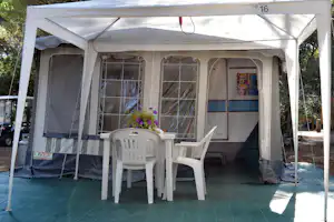 Wohnwagen-Stellplatz unter einem Zeltvorzelt mit weißen Kunststoffstühlen und Tisch auf grünem Boden, seitlich Fenster und Vorhänge.
