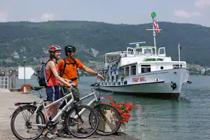 Zwei Radfahrer am Ufer mit Fahrrädern, im Hintergrund ein weißes Ausflugsschiff und bewaldete Hügel.