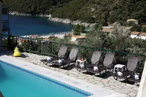 Pool am Campingplatz mit Sonnenliegen und Meerblick, im Hintergrund bewaldete Küste und kleine Häuser am Strand.
