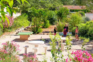 Campinggarten mit Blumenbeeten, Steintischen und Sitzbänken, im Hintergrund Hecken und ein Haus mit Ziegeldach.
