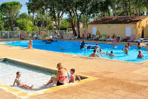 Großer Camping-Pool mit Kindern und Erwachsenen auf Liegen, im Hintergrund Bäume und ein Gebäude mit Terrassenbereich.