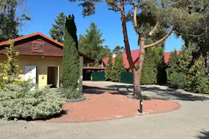 Campinghaus mit Eingangstür, Holzveranda und roten Dächern, gepflasterter Weg und Grünpflanzen im Schatten der Bäume.
