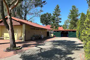 Waschhaus am Campingplatz mit Waschbecken, Waschmaschine und Abfalleimer, gepflasterter Fläche und Bäumen im Vordergrund.