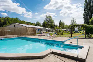 Campingpool mit blauem Wasser, Liegen und Sonnenschirmen, dahinter Gebäude, Hecken und Bäume unter blauem Himmel.