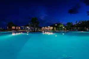 Beleuchteter Campingpool am Abend mit Liegen, Palmen und Gebäuden im Hintergrund.