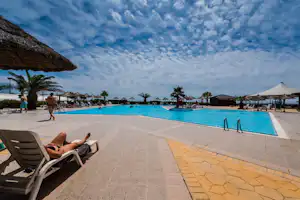 Poolbereich mit Liegen, Sonnenschirmen und Palmen am Campingplatz, im Hintergrund Gebäude und weitere Schirme.