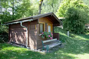 Holzhütte mit Veranda, Bank und Blumenkästen im Grünen, umgeben von Bäumen und Gras am Campingplatz.