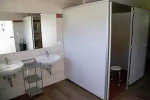 Camping-Badezimmer mit zwei Waschbecken, Duschkabine und Ablagewagen aus Metall, hell geflieste Wände und Boden.